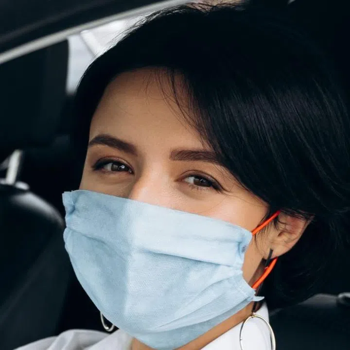 A girl in a protective medical mask sits behind the wheel of her car and looks at the camera