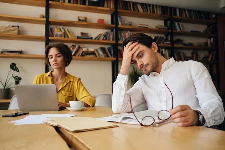 Young tired man sitting at the table with notepad and eyeglasses thoughtfully holding hand near head at work in office with colleague on background