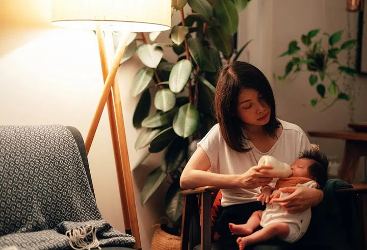 Exhausted  Asian mum feeding infant at night, sitting in the living room.