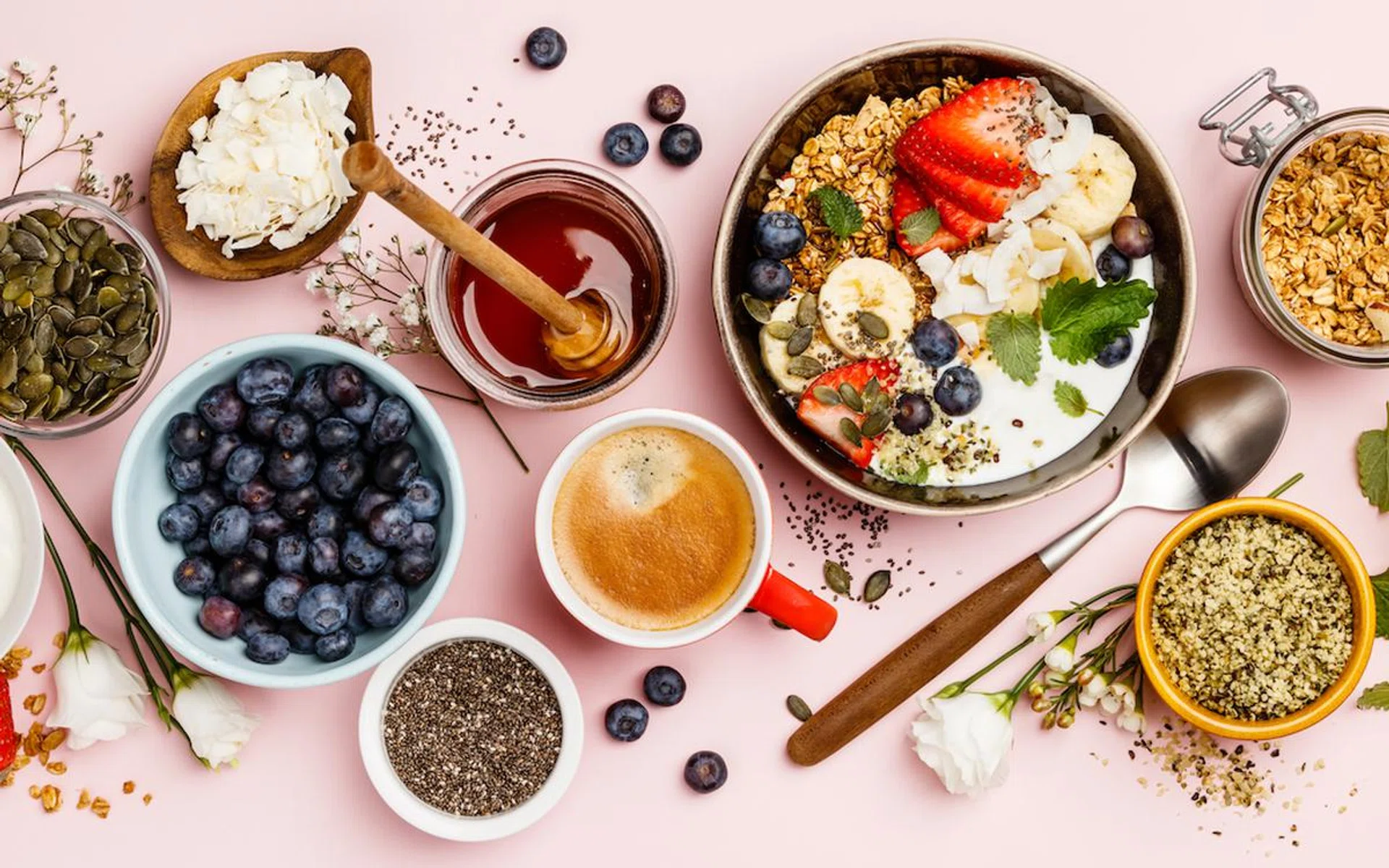 Healthy breakfast set. Oat granola with fresh berries, banana, yogurt, maple syrup, seeds and mint leaves  with cup of coffee on pink background, flat lay, top view