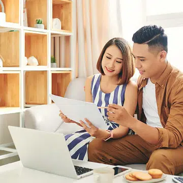 A picture of a happy Asian couple sitting on a sofa, discussing finances, for an article about What Every Married Woman Should Know About Money & How to Get Finances Back On Track