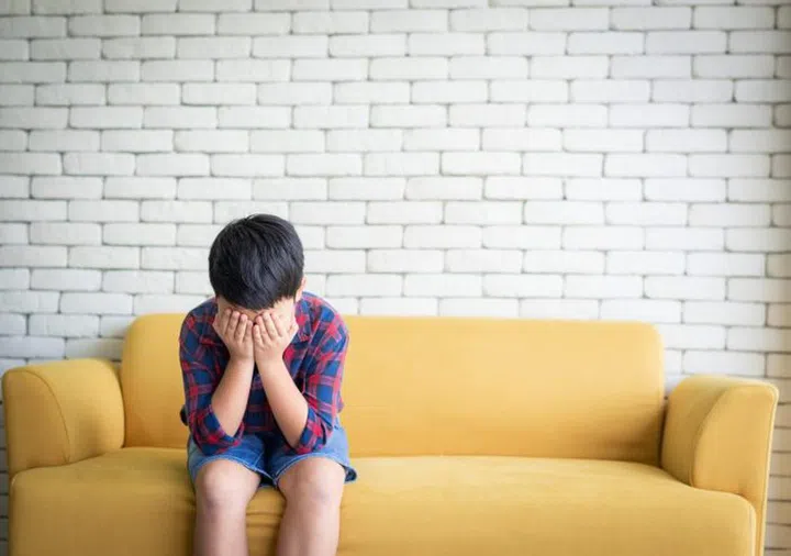Little Asian boy feeling sad sitting alone on sofa and cover his face