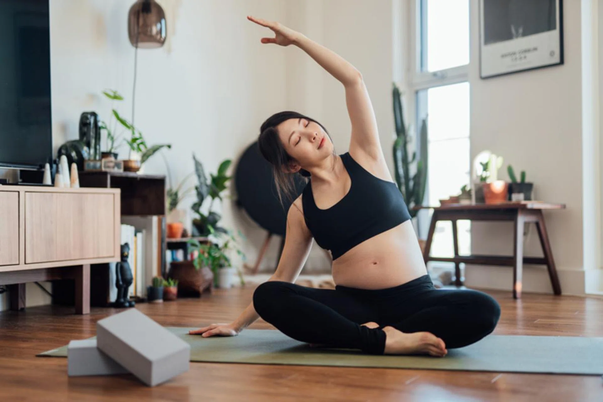 Pregnant Asian woman practices yoga at home in living room. Second trimester. Healthy and active pregnancy lifestyle concept. Revolved Head to Knee Pose - Parivrtta Janu Sirsasana.
