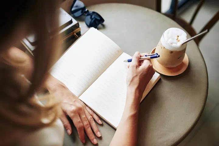 Creative young woman drinking coffee at cafe table and writing in journal her thoughts and ideas
