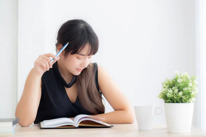 Beautiful asian woman writer smiling thinking idea and writing on notebook or diary with happy on desk office