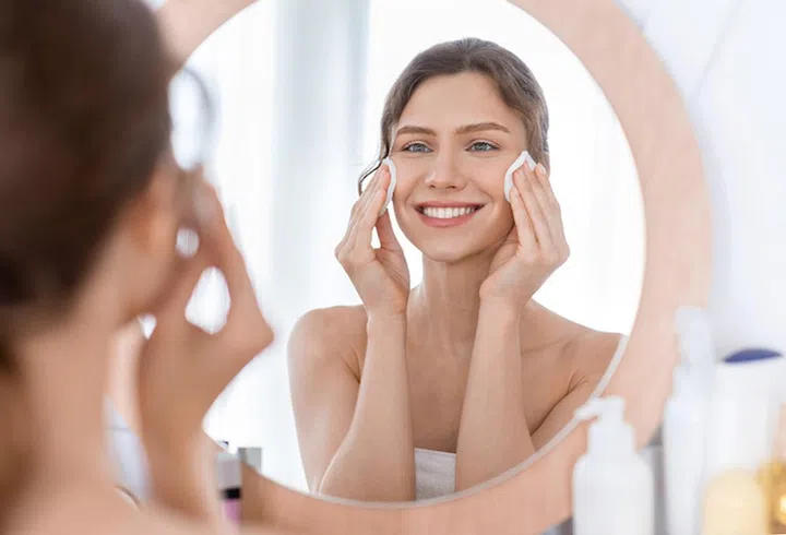 Skincare. Young cheerful woman looking at mirror and cleaning her face with cotton pads at home