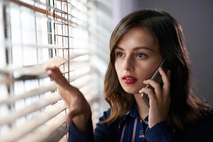 Beautiful young business lady talking on phone and looking through blinds