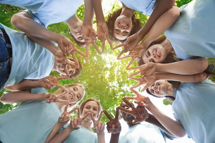 Group of happy volunteers standing in circle in park and showing peace or v sign, view from below, copy space