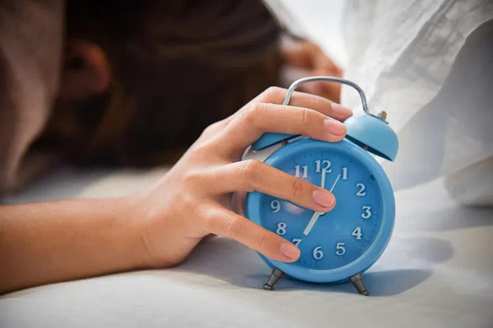 Sleepy Asian woman extending hand to Alarm clock on her bed. Female having trouble waking up. Stress from overtime working concept. Nightmare.