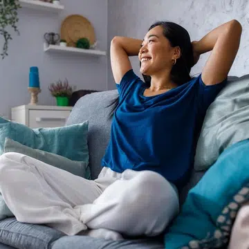 Full length shot of beautiful, happy young woman sitting on the cozy sofa with hands behind head, looking away, smiling and daydreaming.