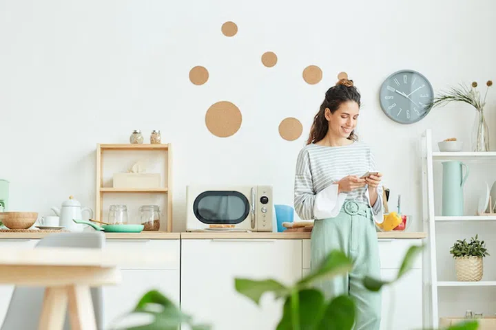 Happy young woman wearing stylish outfit standing in her kitchen texting message to someone using her smartphone