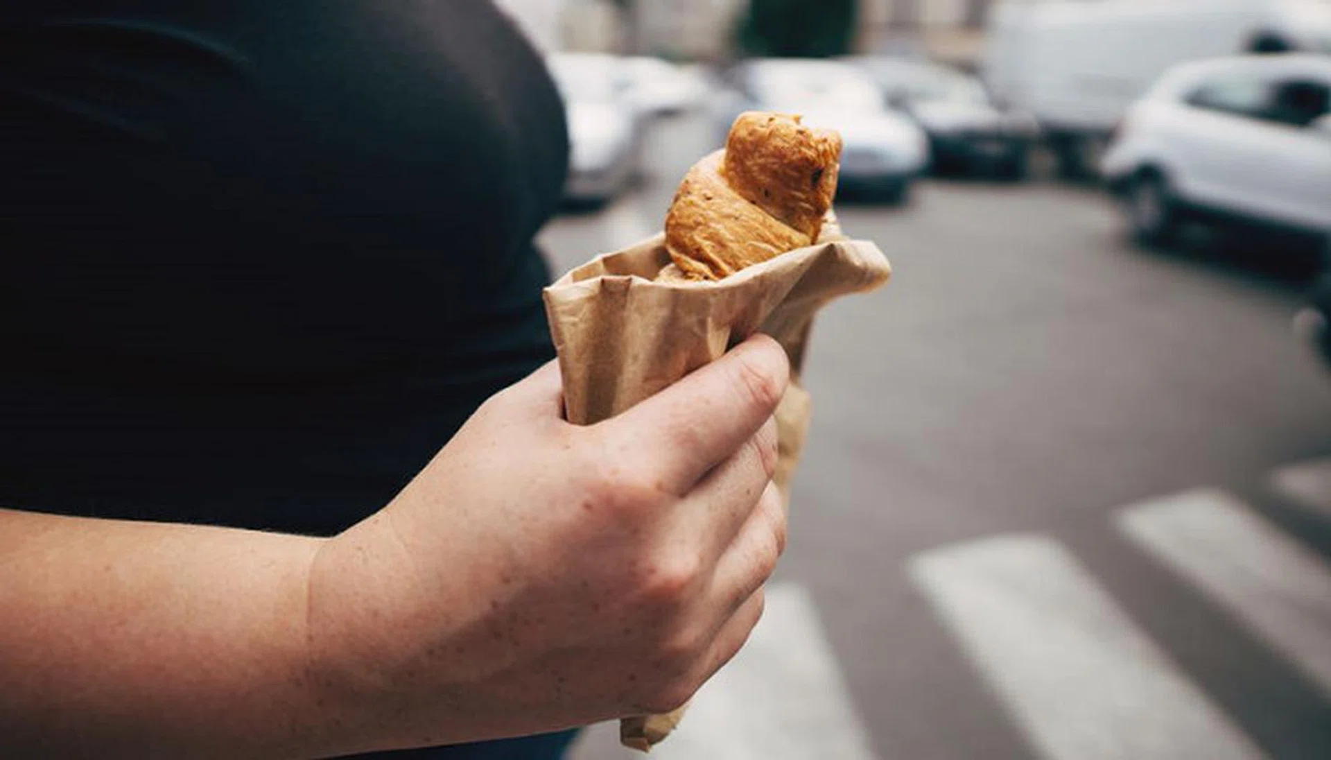 Unhealthy snack, eating on the go, junk food, overweight concept. Obese woman woman eating croissant in paper bag walking in the street