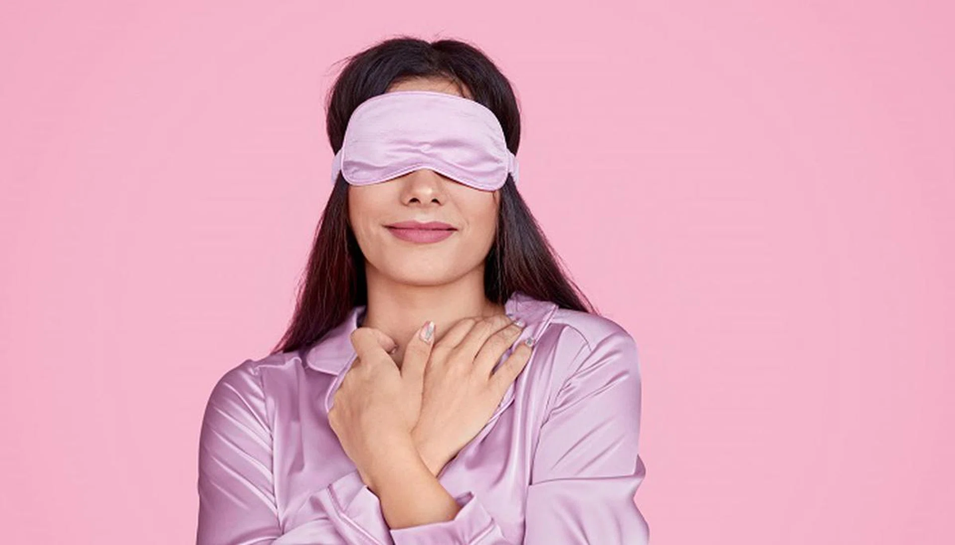 Young lady in sleep mask touching delicate silk sleepwear and enjoying softness and comfort while resting against pink background