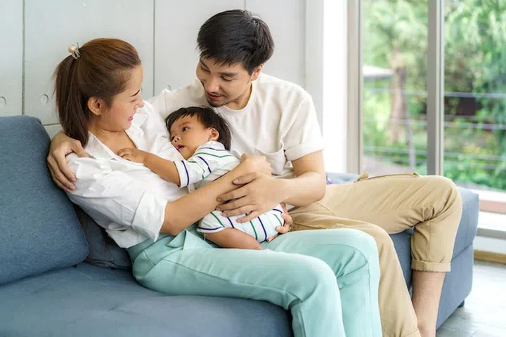 Asian father and mother are hugging 9 months baby son on sofa. They are smiling and warm touching to the baby with love in living room at home