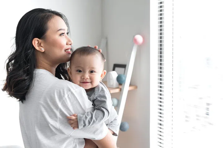 Beautiful young mom is playing with her cute baby and smiling while standing near the window at home