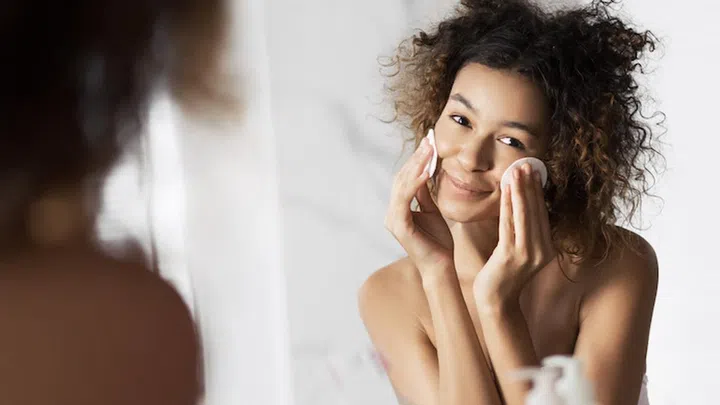 Beautiful afro-american girl cleaning face with cotton pads looking in mirror at bathroom. All skin types cleansing concept
