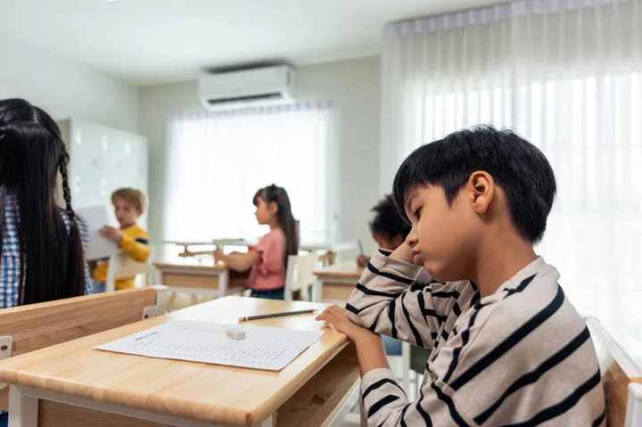 Asian young boy student doing an exam test at elementary school. Adorable children sitting indoors on table, feeling upset and depressed while writing notes, learning with teacher at kindergarten.