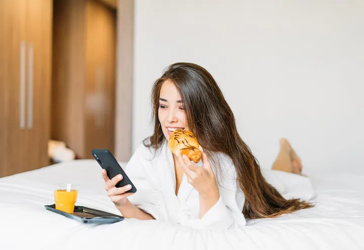 Beautiful young asian woman with long hair in white robe using mobile having breakfast croissant and orange juice in bed of hotel room