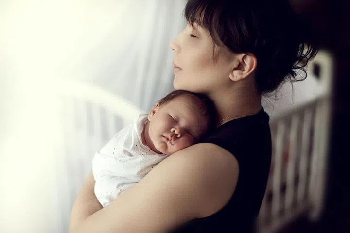 young Asian mother tenderly holds the sleeping on the shoulder of the baby in the nursery on the background of the crib
