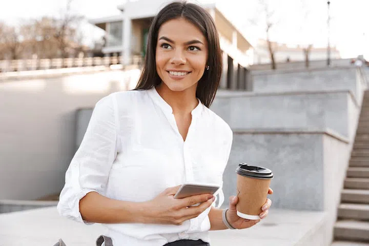 Smiling brunette woman in shirt sitting outdoors and looking away while holding smartphone and cup of coffee