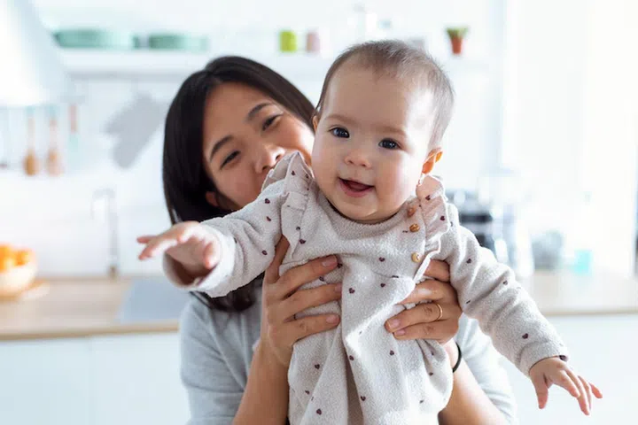 Shot of happy young mother with her baby daughter looking at camera in the kitchen at home.