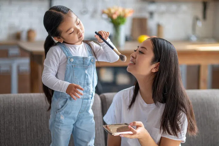 Mother seated on the sofa holding the powder-box in her hand while her daughter powdering her face