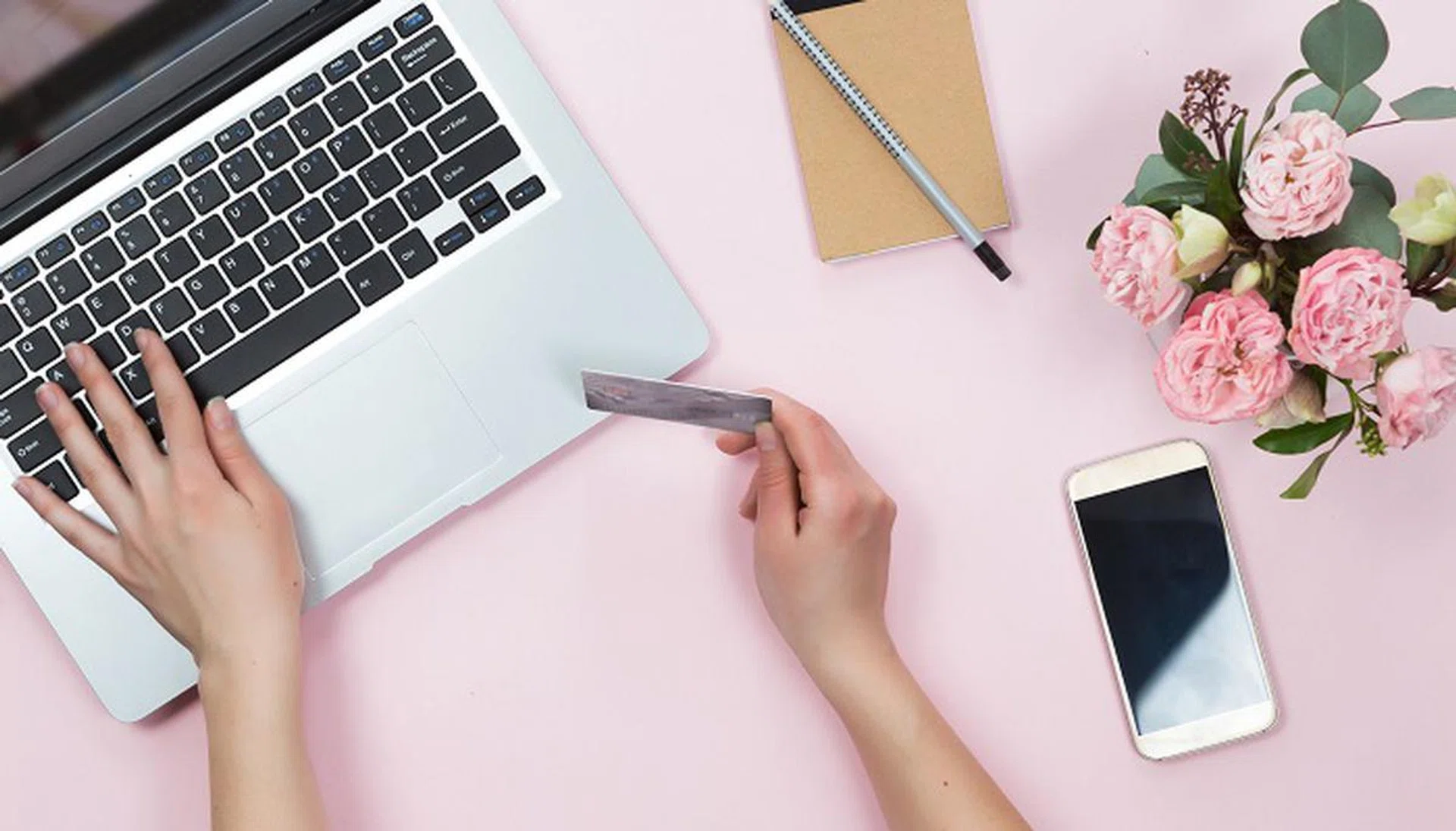 Top view of woman hands holding credit card, online shopping concept, workspace with laptop, mobile phone, flowers and notebook, flat lay.