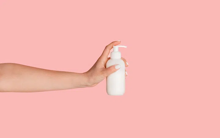 Closeup of female hand showing dispenser bottle with cosmetic product over pink background. Panorama