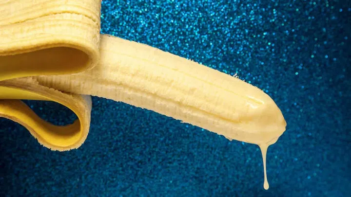 Condensed milk drips from the end of a banana on shiny blue background. The concept of ejaculation, emission or sperm. The peel of a yellow banana is collected in the form of male testicles.