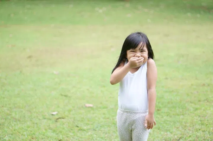 Asian children cute or kid girl stand smile with laugh and hand close mouth for happy fun with enjoy or gag and secrets on green grass meadow space with wear white vest