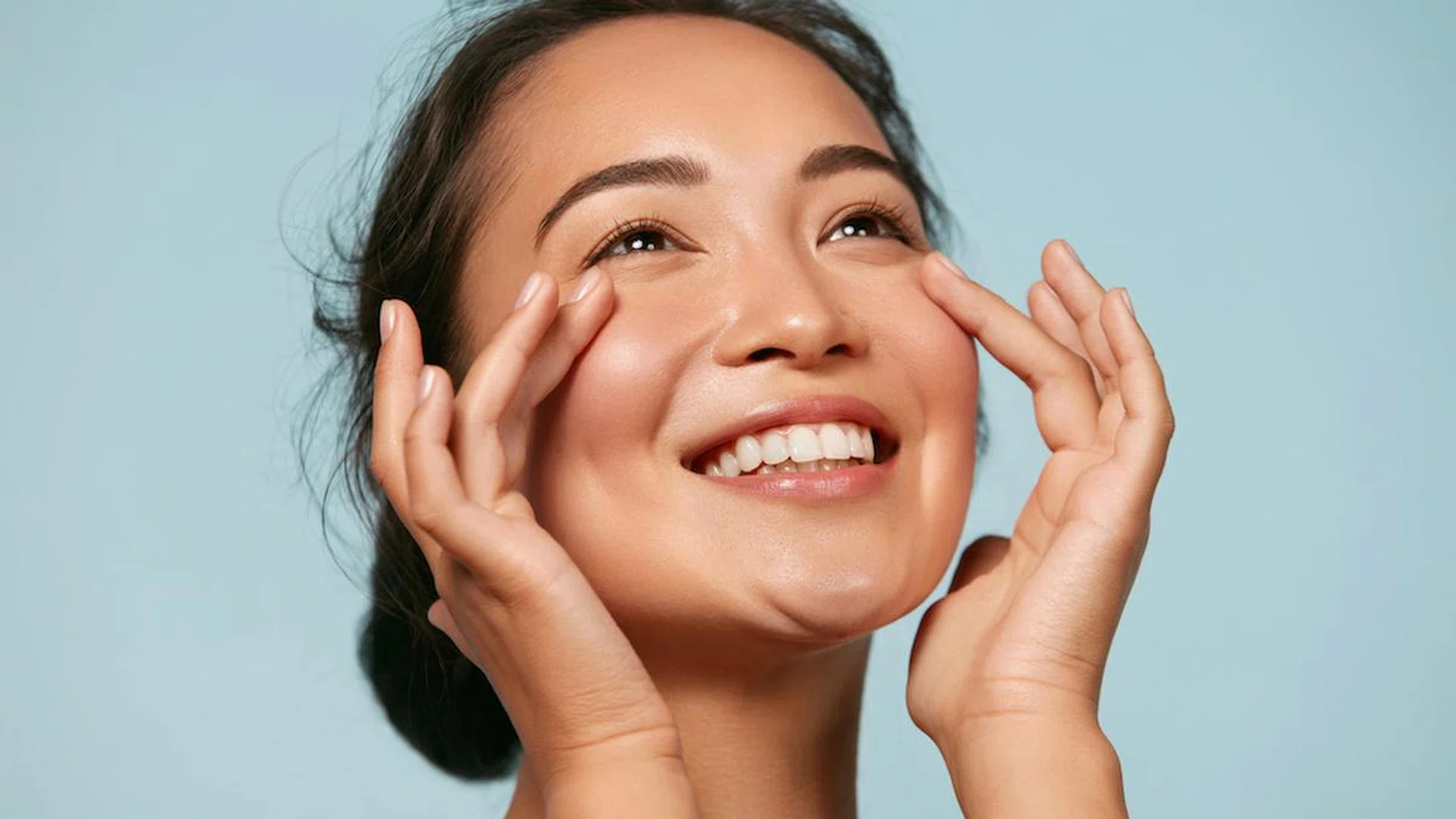 Skin care. Woman with beauty face touching healthy facial skin portrait. Beautiful smiling asian girl model with natural makeup touching glowing hydrated skin on blue background closeup (Skin care. Woman with beauty face touching healthy facial skin p