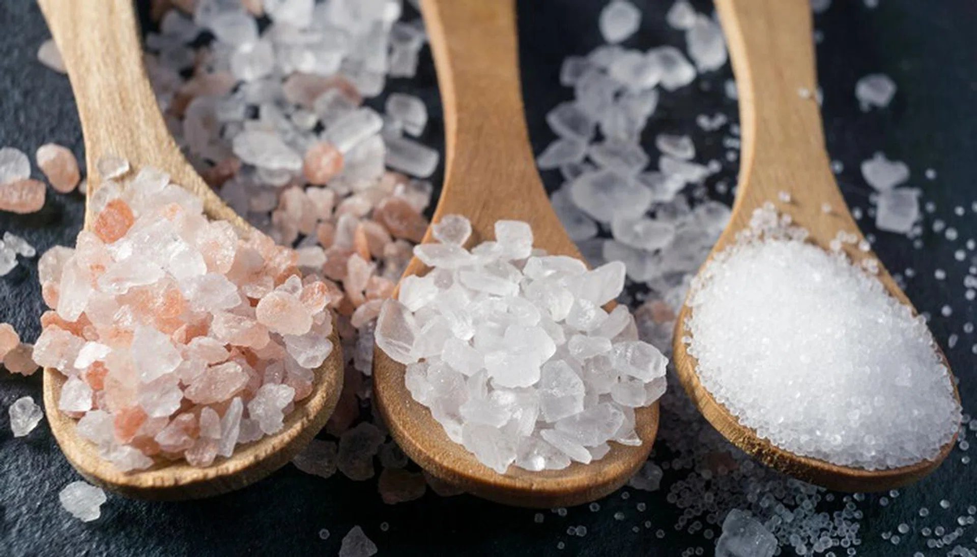 Different types of salt. Sea, himalayan and kitchen salt. Top view on three wooden spoons on black background