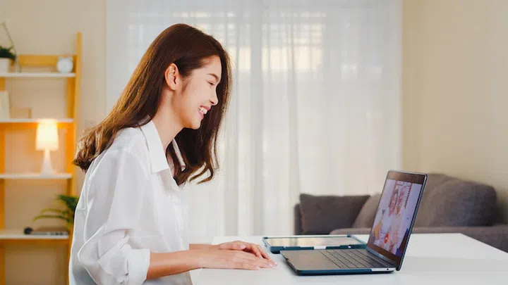 Young Asian business female using laptop video call talking with family dad and mom while working from home at living room. Self-isolation, social distancing, quarantine for coronavirus prevention.