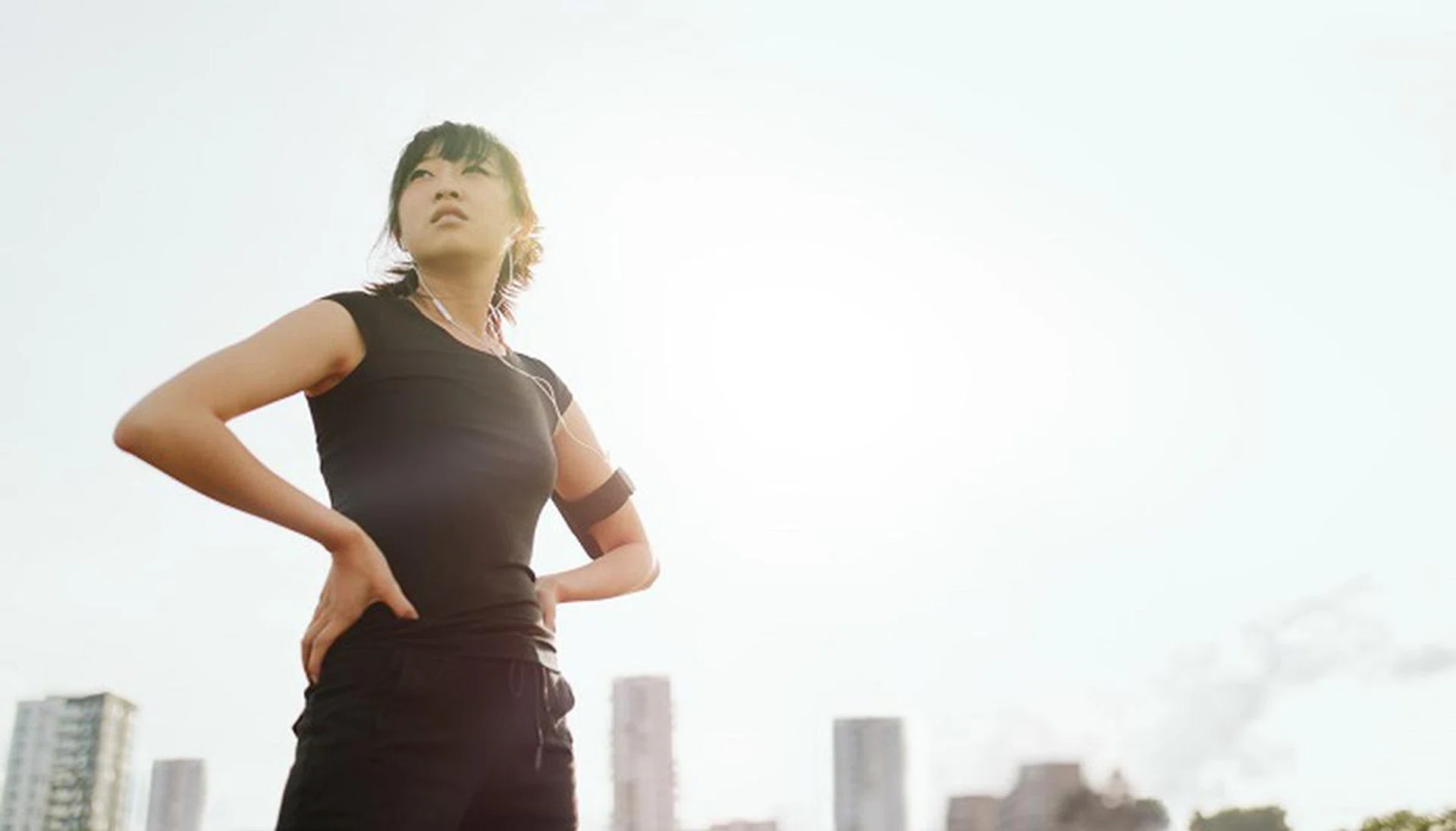 Outdoor shot of fitness woman standing with her hands on hips and looking away. Female runner taking break from outdoor training in morning.