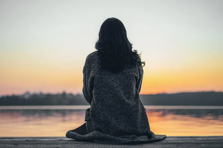 Back view of long hair brunette girl sitting on the pier near lake at sunset time