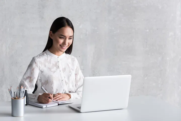 Happy businesswoman is sitting near workplace and making notes. She looking at screen of laptop with smile. Portrait. Copy space on right side