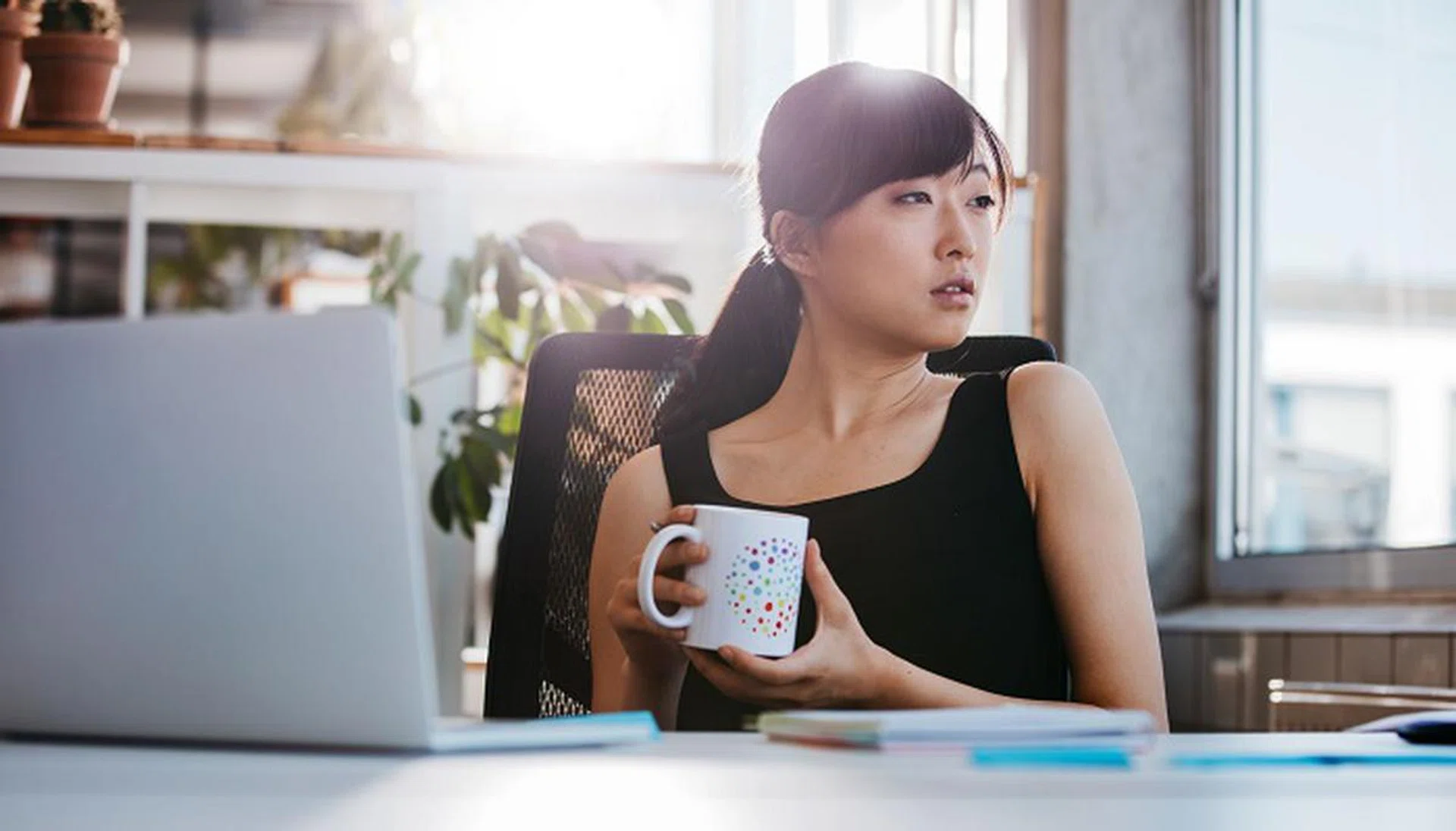 Portrait of relaxed young woman sitting at her desk holding cup of coffee and looking away. Asian business woman taking coffee break in office.