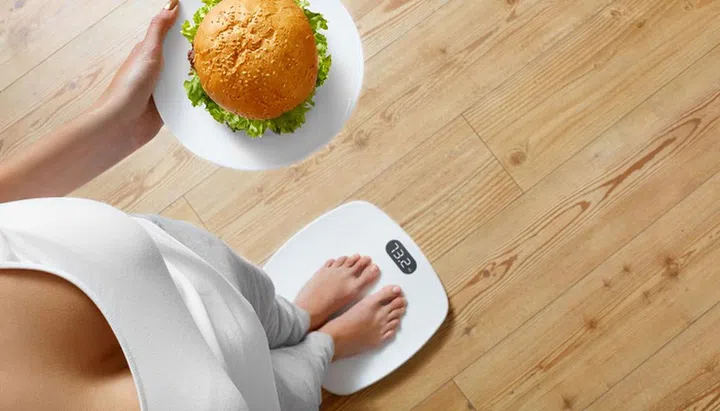 Diet And Fast Food Concept. Overweight Woman Standing On Weighing Scale Holding Burger ( Hamburger ). Unhealthy Junk Food. Dieting, Lifestyle. Weight Loss. Obesity. Top View