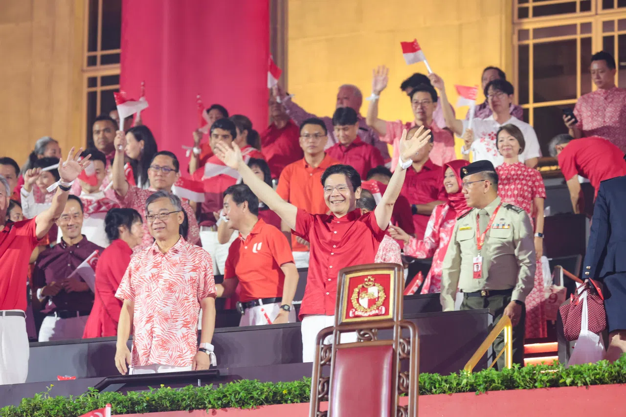 今年5月15日就任后，首次以总理身份参加国庆庆典的黄循财（前排中）乘专车抵达现场后，在观众的热烈掌声中登上观礼台挥手致意。在他身旁的是同样在5月升任副总理兼贸工部长的颜金勇（左）。（李冠卫摄）