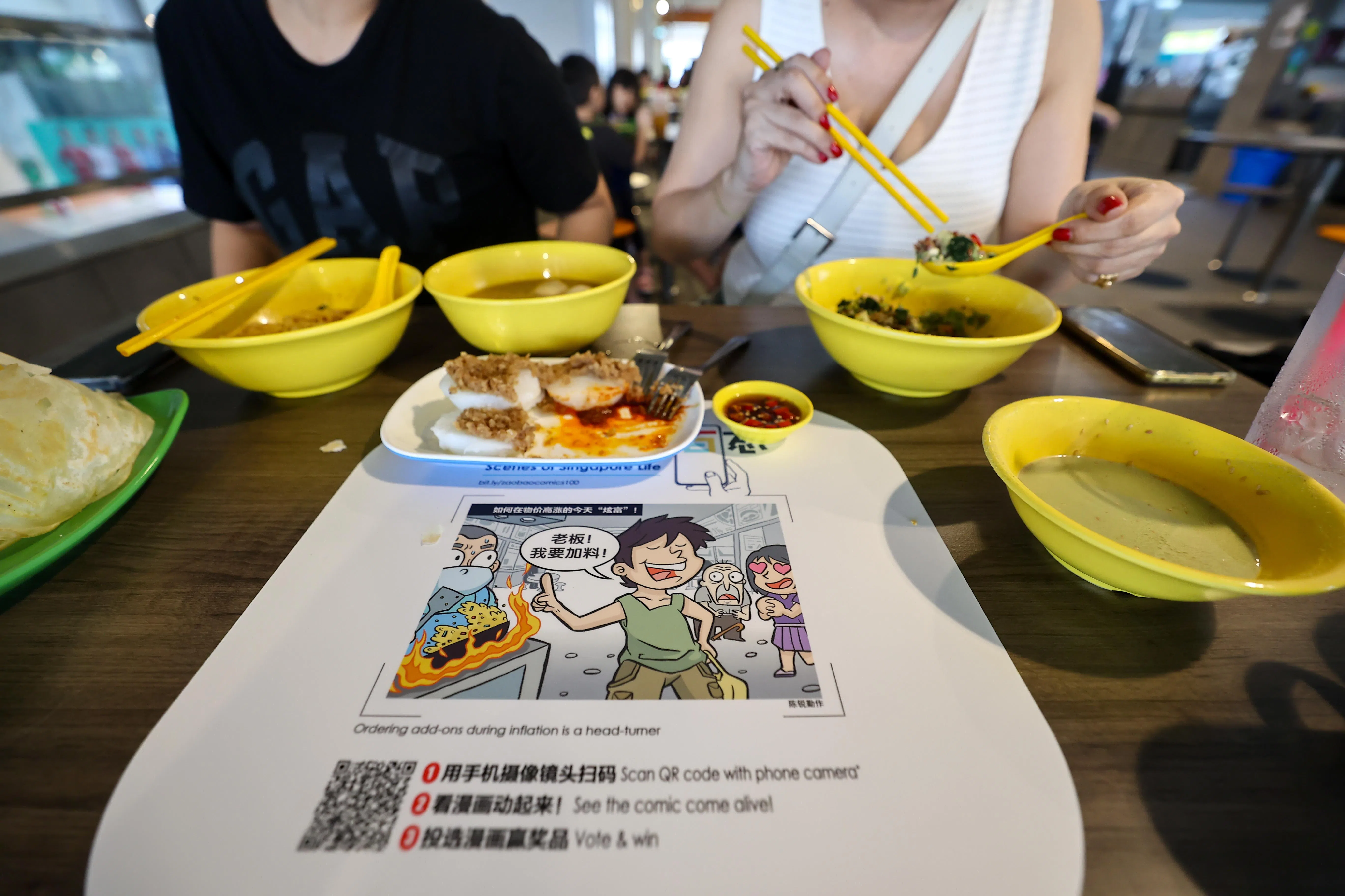 10座小贩中心的桌面贴有漫画贴纸，用手机摄像头扫描QR码，还能这些漫画跃然纸上动起来。（陈渊庄摄）