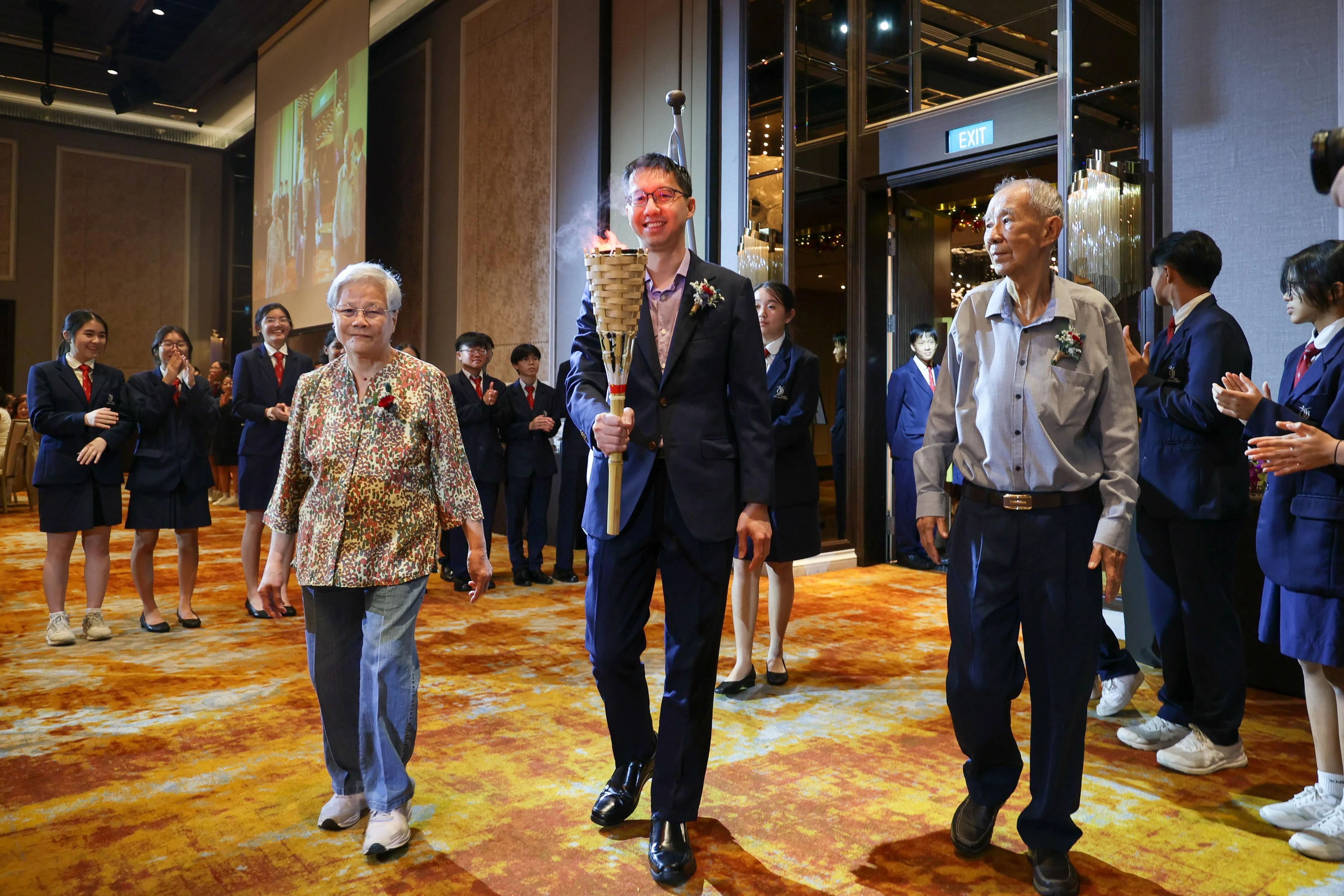 新民中学校长陈顺惠（中）手持“新民传承跑”火炬步入宴会厅，两旁分别是校友叶小琳（左）以及黄安嵘（右）。叶小琳是学校创始人叶帆风的女儿。（张俊杰摄）