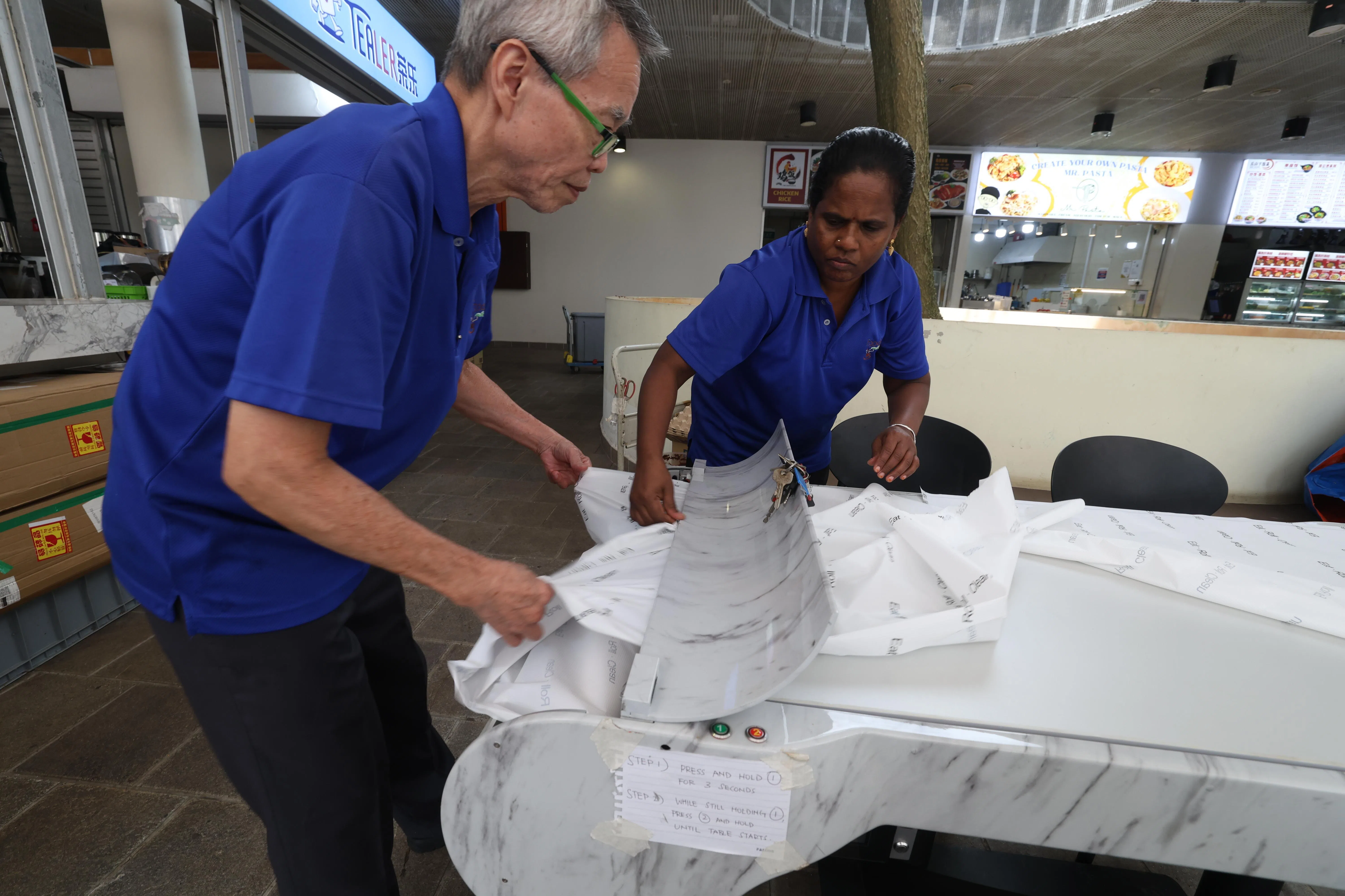 南大食阁工作人员李阿华（左）和瓦桑塔，示范如何为自动清洁餐桌进行维护，他们正在将用过后卷起来的桌布取出来。（关俊威摄）