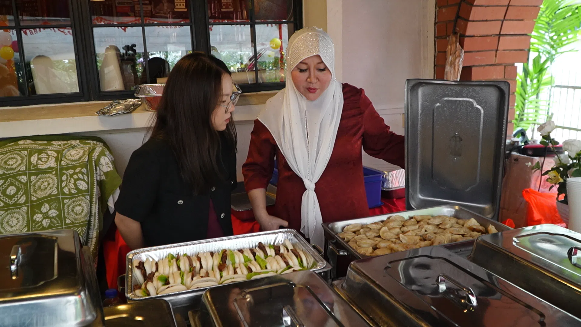 来自中国的回族新移民海丽娟（右）向记者张千雪（左）介绍新春聚会的食物。她做了大盘鸡、酱牛肉和水饺等回族特色的食物，给本地华族回教徒品尝。（视频截图）