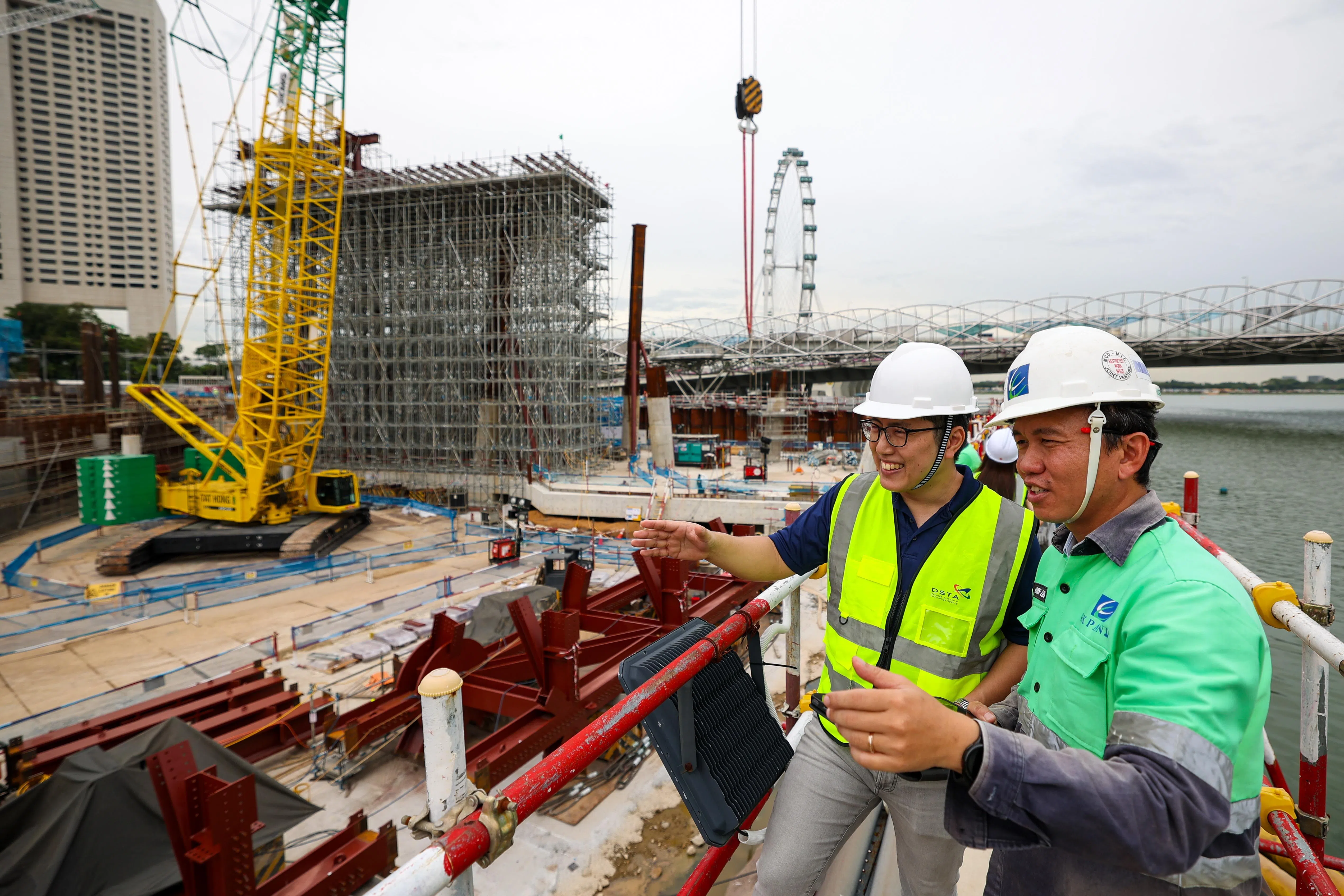 国防科技局高级项目经理黄树恩（左）和Expand建筑公司项目部助理主任林子洋，从“浮动办公室”出发，经过连接通道，步行两三分钟到卫国广场工地进行视察。图中黄色塔吊旁被金属脚手架裹得严严实实的建筑是卫国广场空中泳池，建成后将与金沙酒店顶部的无边泳池，隔岸相望，相映成趣。（林明顺摄）