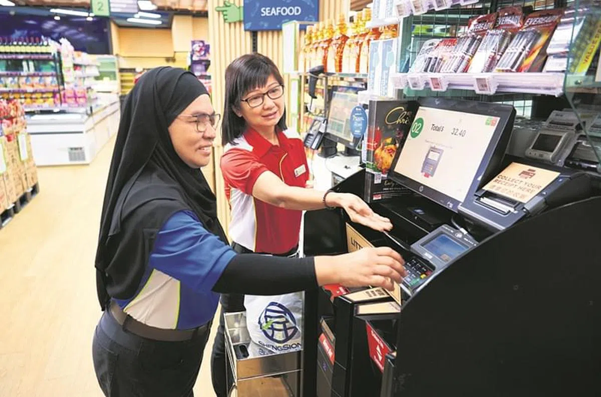 吴秀华（红衣者）教导新收银员罗娜妮使用混合式自助收款系统，系统操作容易，她很快就上手。（图/新报业媒体）