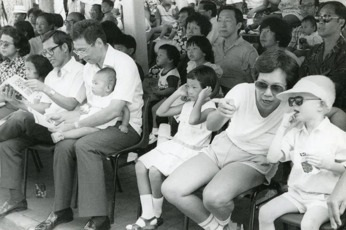 李显龙（抱着鸿毅）与何晶（右二）婚后，何晶为他再添两个儿子，图为夫妇带着原配所生的修齐和毅鹏到乌节路看妆艺大游行。坐在李显龙右手边的是前内阁部长李玉全。（档案照片）