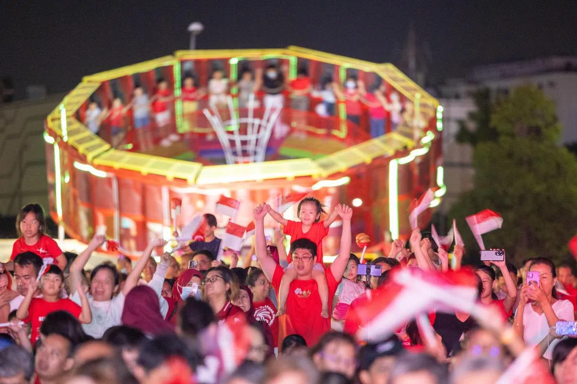 暮色降临，在义顺参加国庆邻里嘉年华的公众边看舞台表演，边哼唱国庆主题曲和挥着红白星月国旗，沉浸在国庆的氛围中。（吴先邦摄）