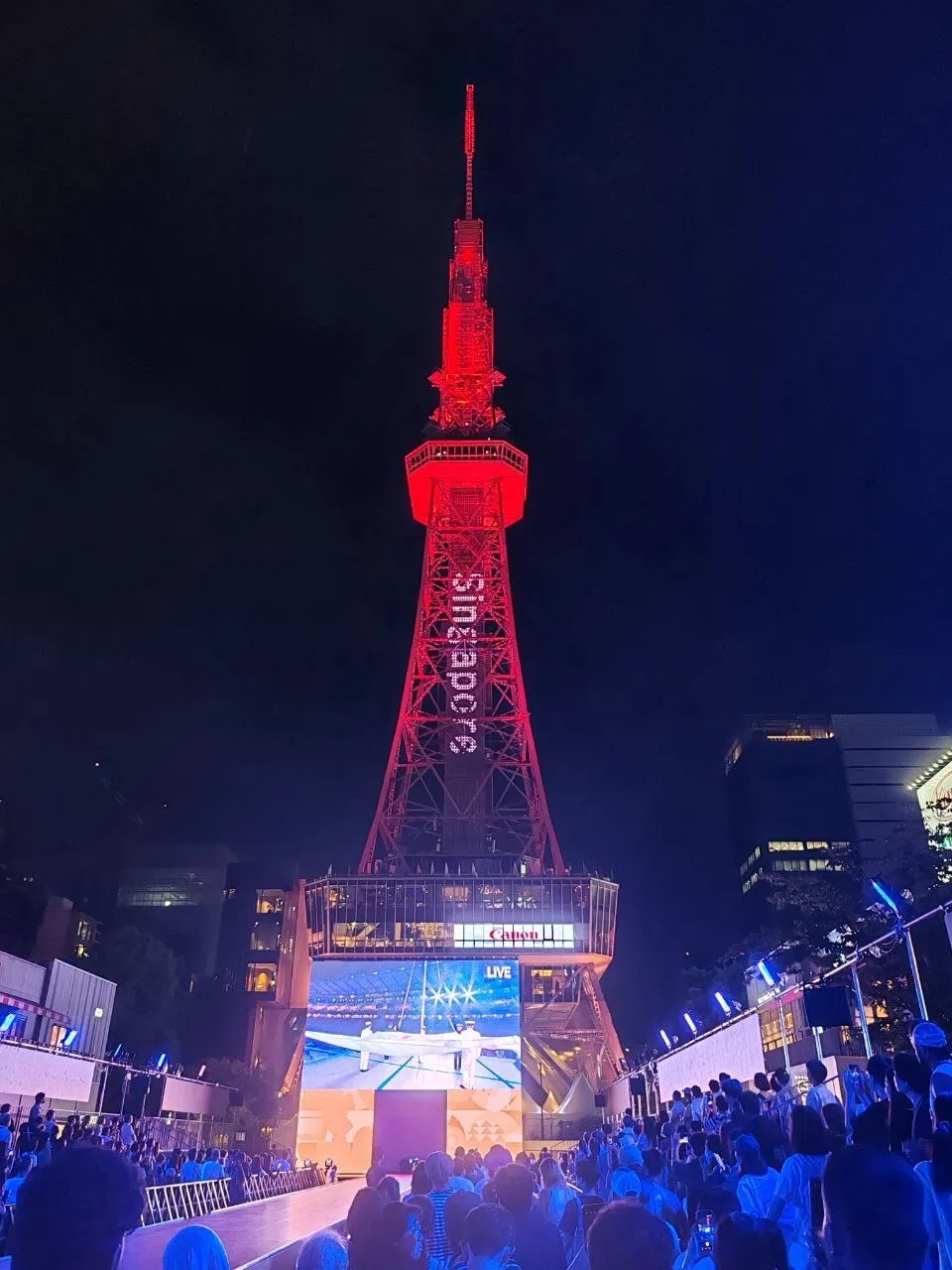 名古屋地标中部电力塔轮流展现各国名称和旗帜，图为“新加坡”出现在塔上。现场也举行嘉年华活动，不少公众在现场观看开幕式直播。（陈明耀摄）
