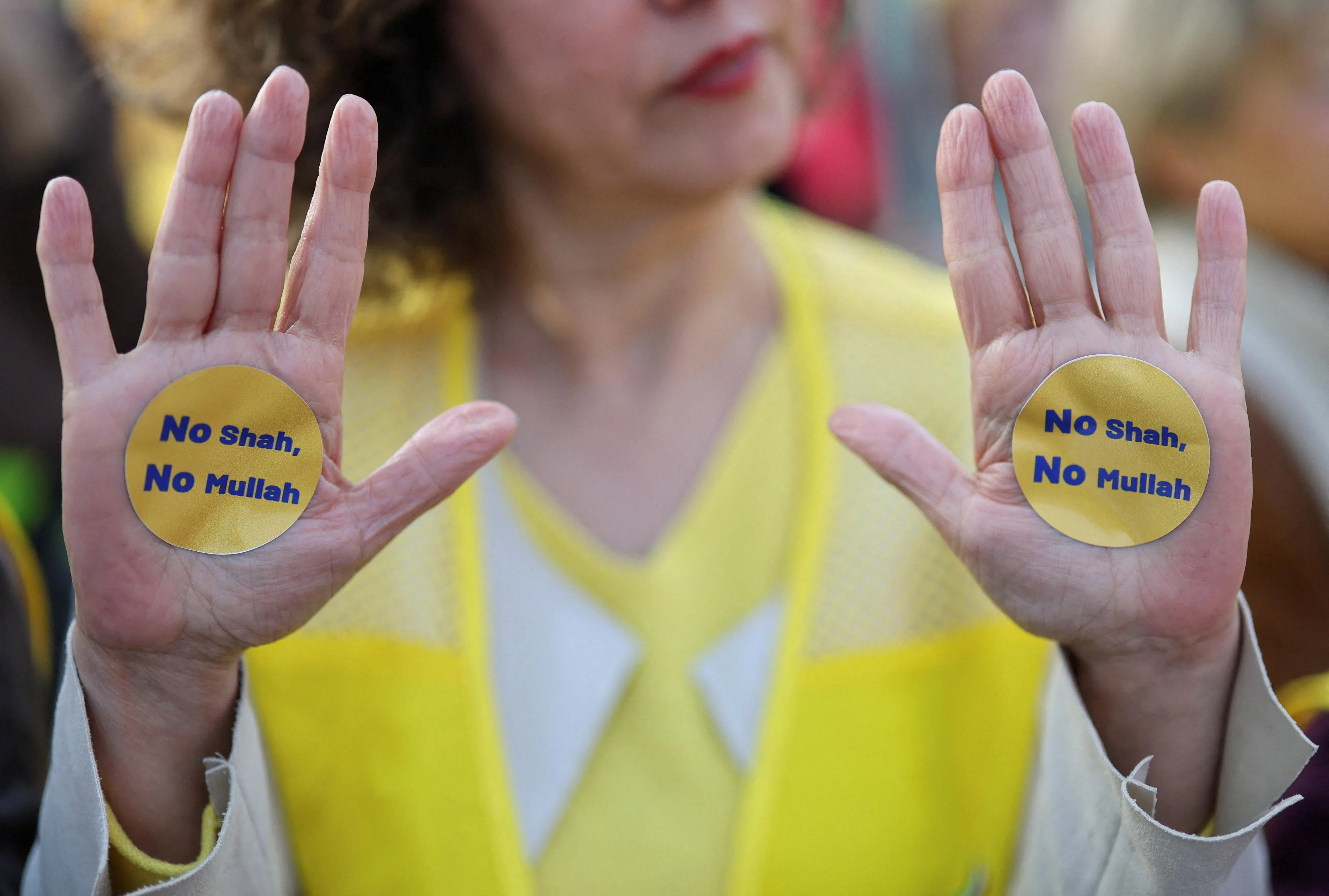 身穿印有“自由伊朗”字样黄色背心的抗议者展示手上贴着的贴纸，上面写着“不要国王，不要毛拉”。“毛拉”指的是伊朗的宗教领袖或神职人员。（路透社）