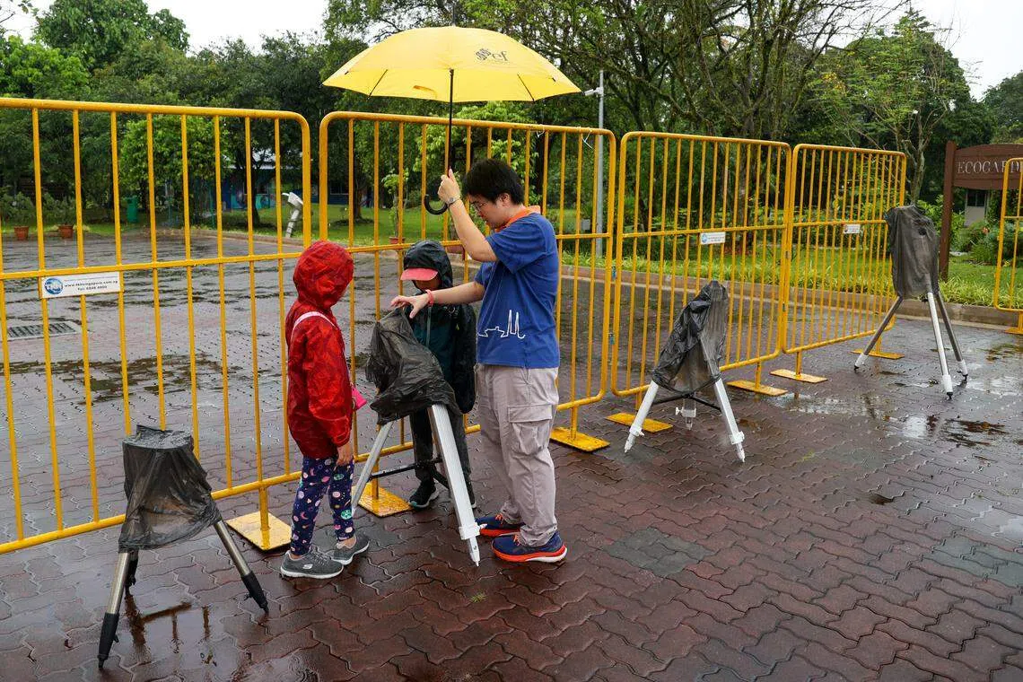 由于新加坡多地阴雨连绵，本地公众今天无缘观测罕见的混合日食。图为科学中心职员为两名孩童解释望远镜需固定在地上，才可观测日食。（蔡家增摄）